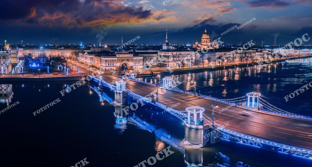 На фотографии изображён Дворцовый мост, освещённый яркой иллюминацией в ночное время. На заднем плане выделяются Исаакиевский собор и здания Адмиралтейства, подчёркивая историческую значимость этой части города. Отражения огней в водах Невы добавляют снимку атмосферу зимней сказки. Ночной Санкт-Петербург раскрывает свою красоту.